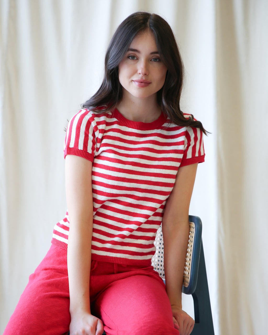 Woman wearing a red and white striped shirt and pink pants sitting against a white curtain.